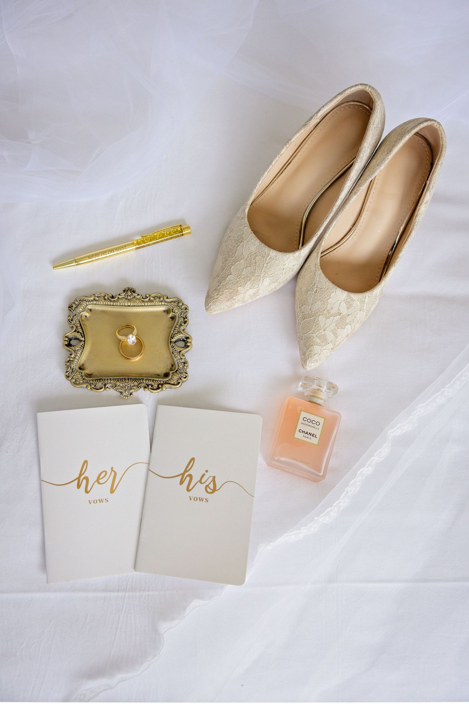 Elegant wedding accessories including white heels, golden frame, perfume bottle and invitation cards on white fabric.