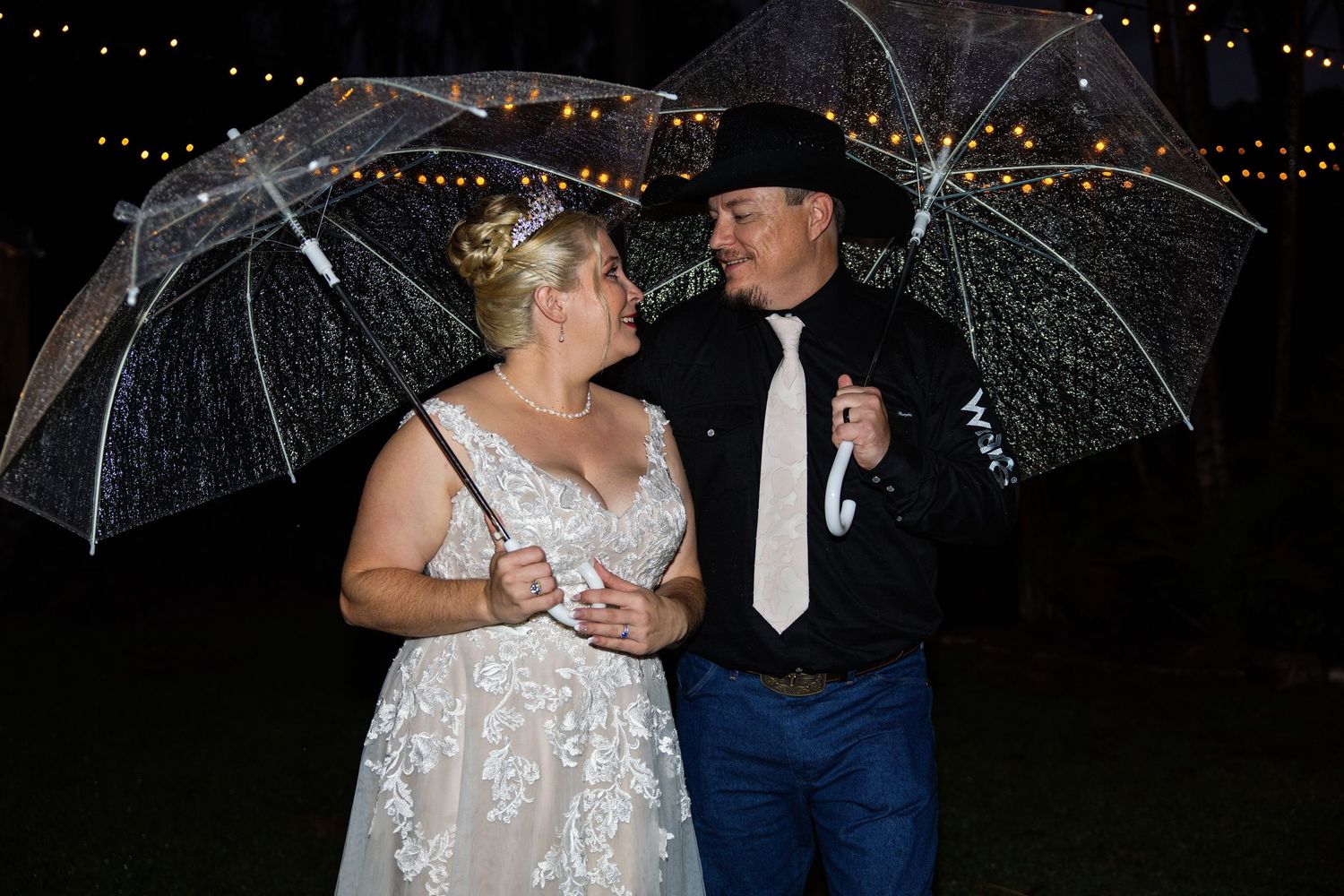 Bride and groom sharing a romantic moment under clear umbrellas in the rain at their outdoor evening wedding reception.