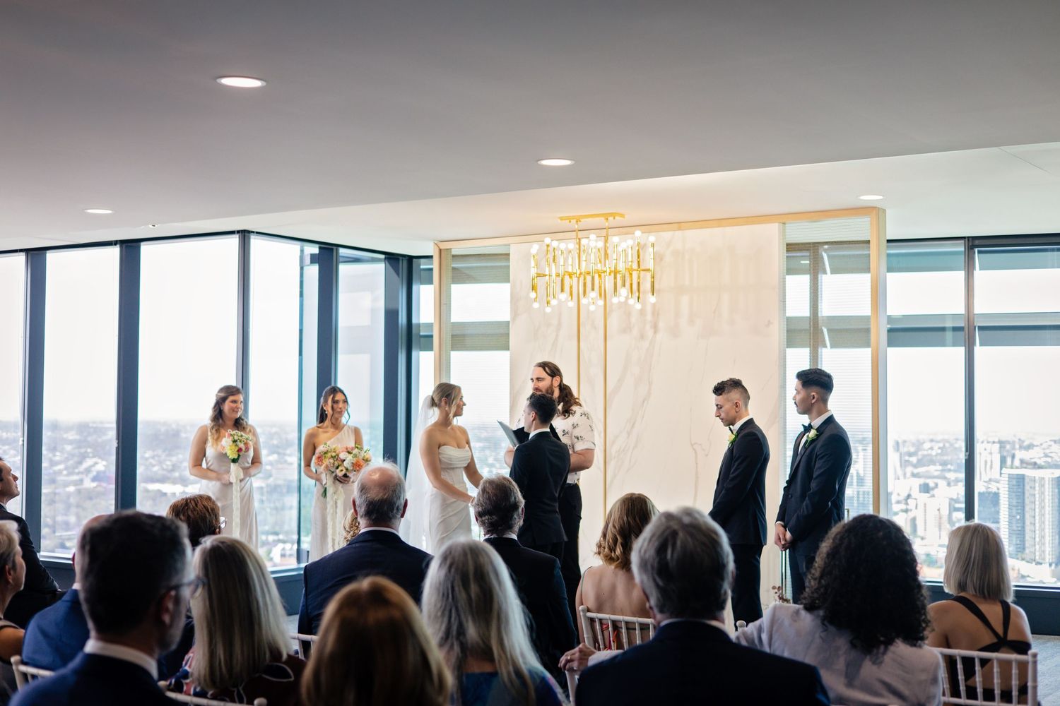Wedding ceremony taking place in modern venue with floor-to-ceiling windows and guests seated in rows.