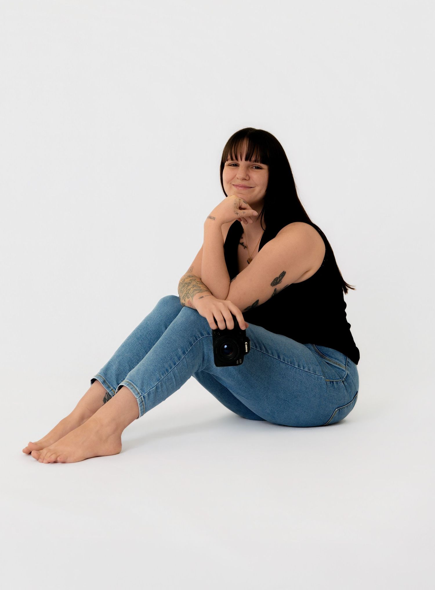 Young woman with dark hair sitting on floor in black tank top and blue jeans, holding phone, smiling at camera.