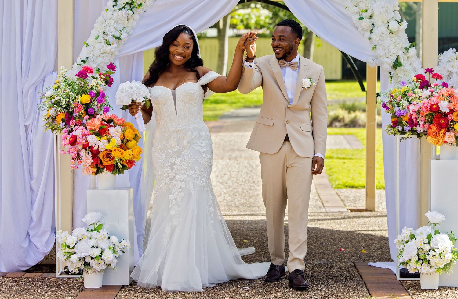 Joyful newlywed couple celebrating under floral arch, bride in white gown, groom in tan suit, colorful bouquets.