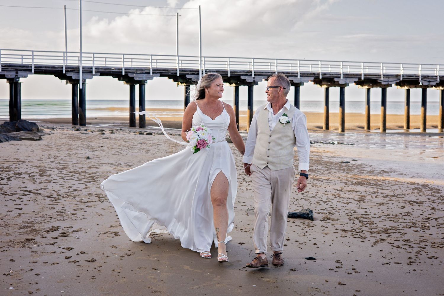 Couple in white attire walking along a beach boardwalk during a seaside photoshoot.