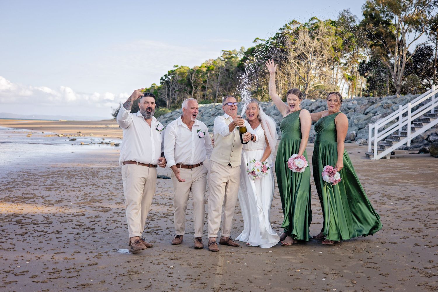 Wedding party celebrating on a beach, bride in white gown, bridesmaids in green dresses, groomsmen in beige suits.