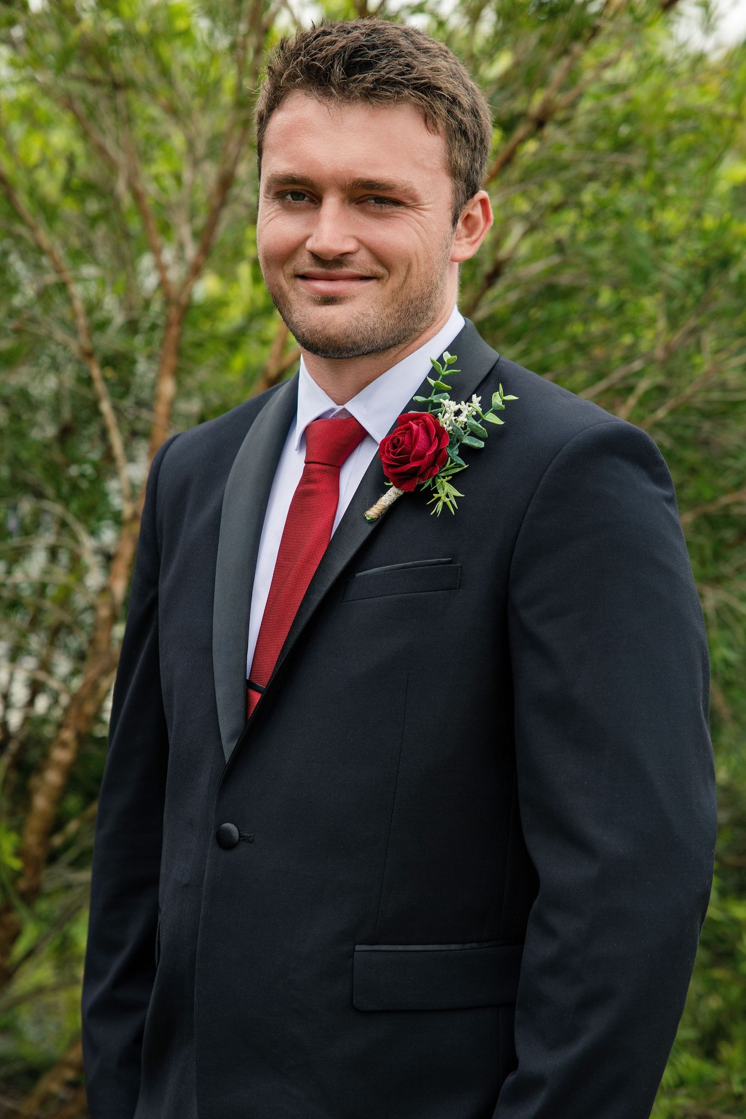 A groom in a black suit with red tie and boutonniere poses outdoors against a natural green background.
