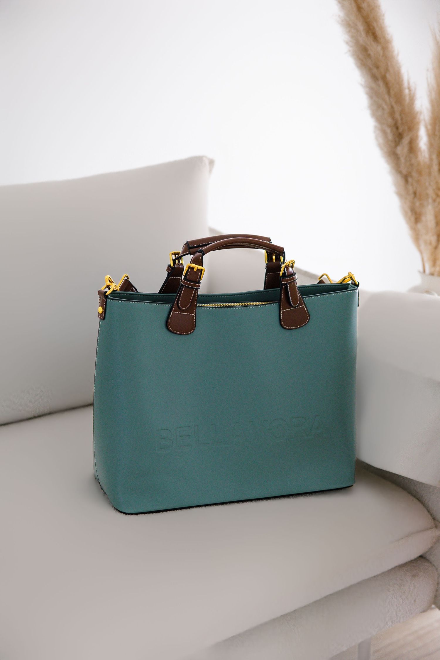 Teal leather tote bag with brown handles and gold hardware sitting on a white sofa.