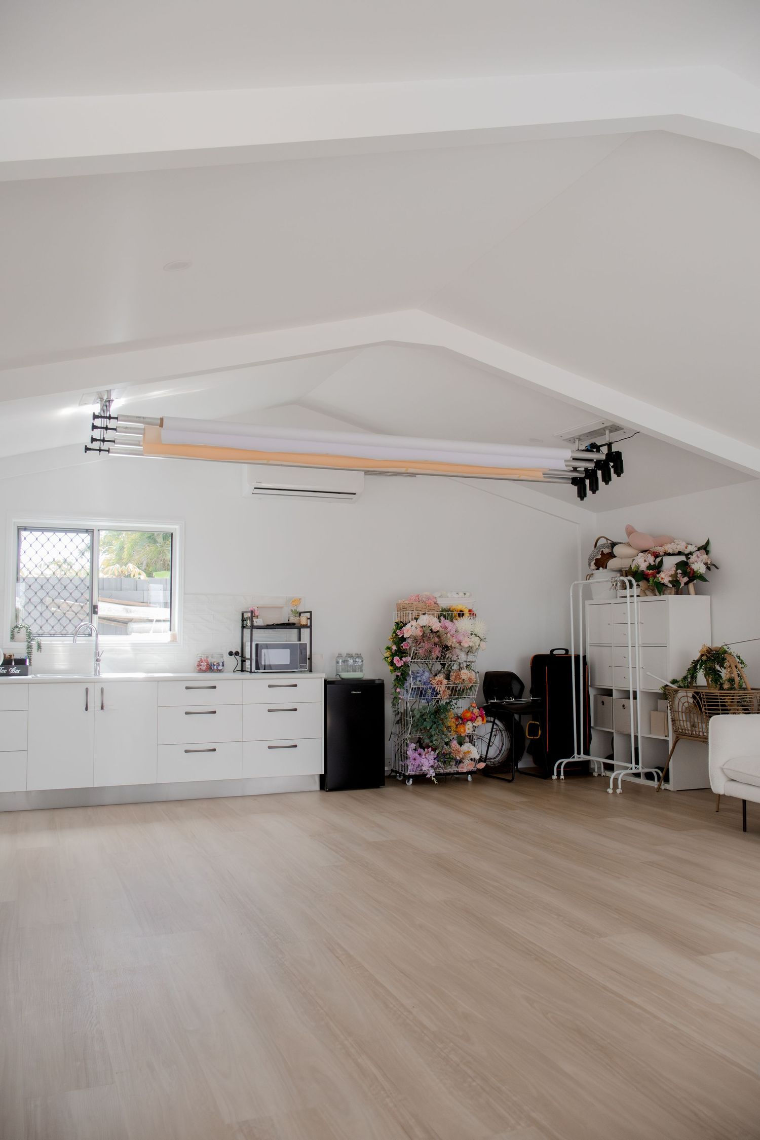 Bright white studio space with vaulted ceiling, hardwood floors, kitchenette, floral arrangements, and photography lighting equipment.