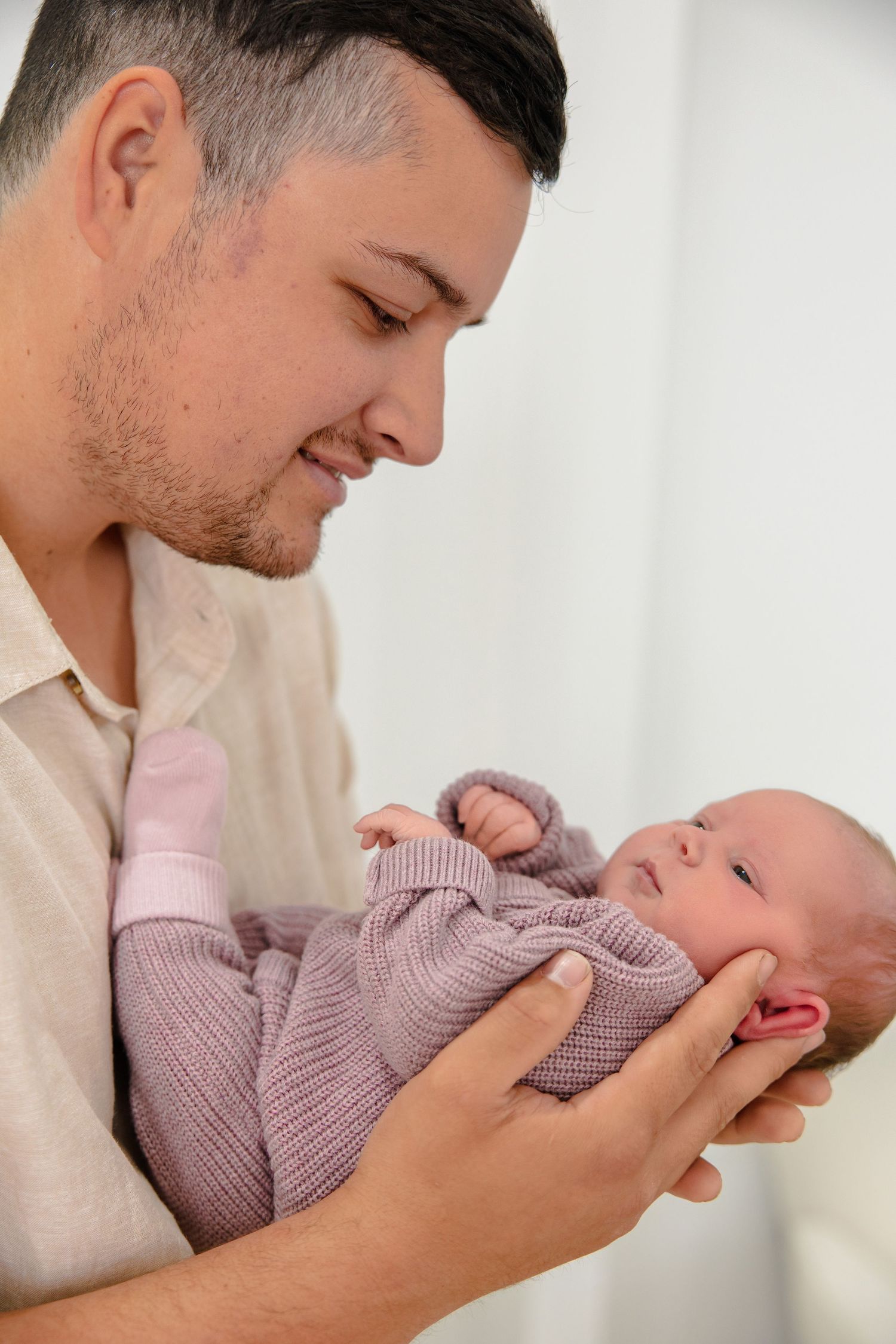 A young father tenderly holds his newborn baby dressed in a purple knit outfit, smiling down lovingly.