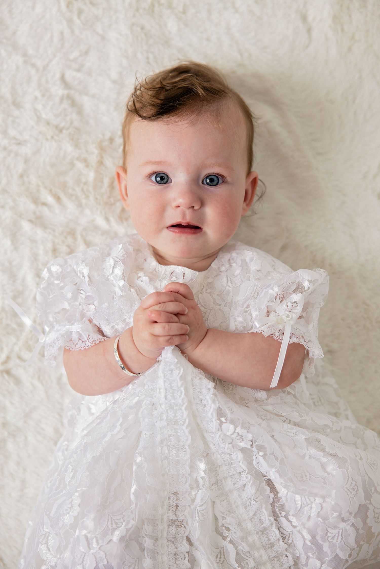 A baby in a white lace christening gown lies on a cream-colored blanket in a series of sweet portraits.