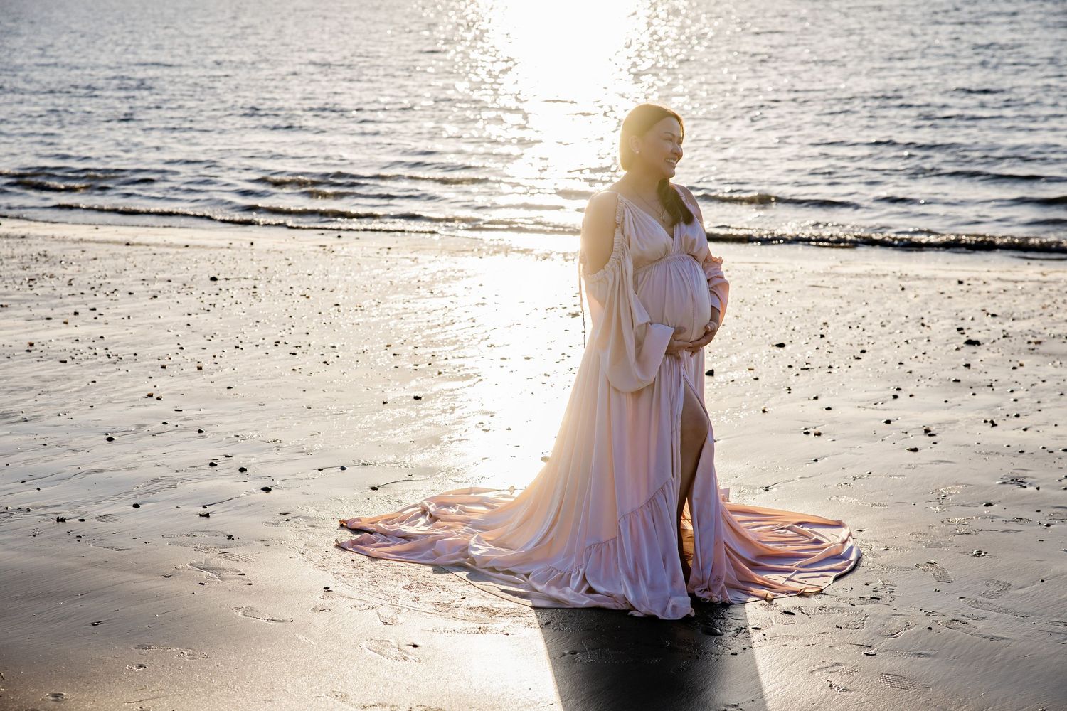 Pregnant woman in flowing pink gown poses on sunlit beach at golden hour for maternity photo.