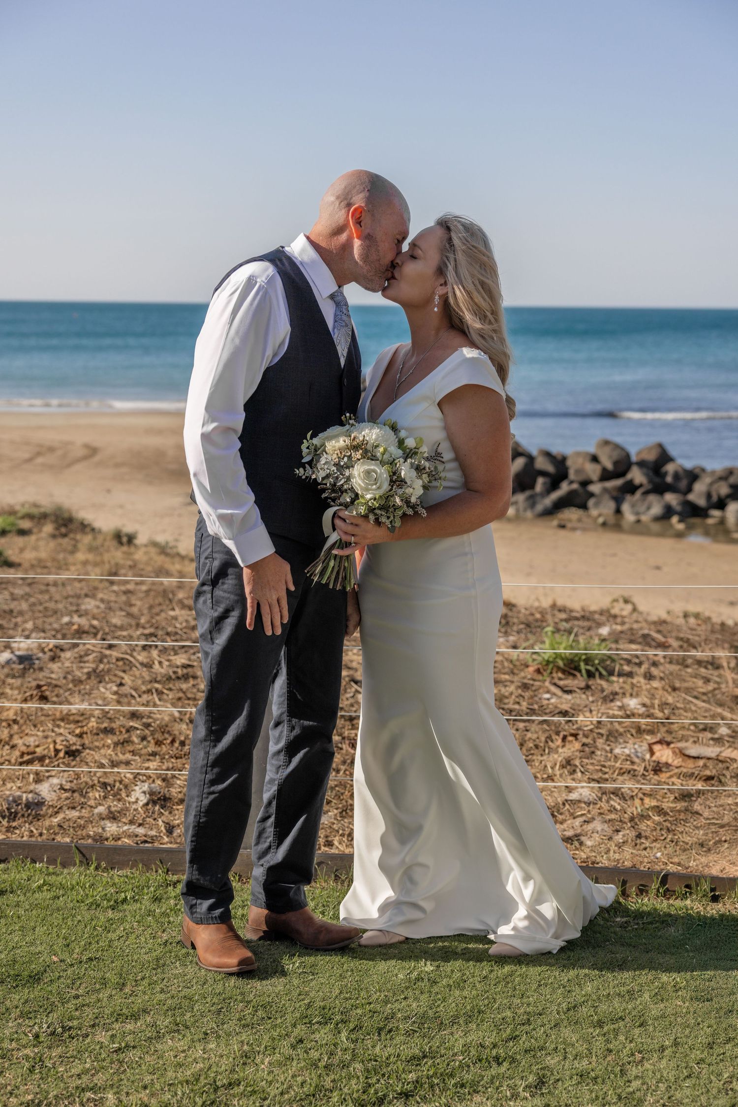 A couple in wedding attire shares an intimate moment on a grassy area overlooking a scenic beach coastline.