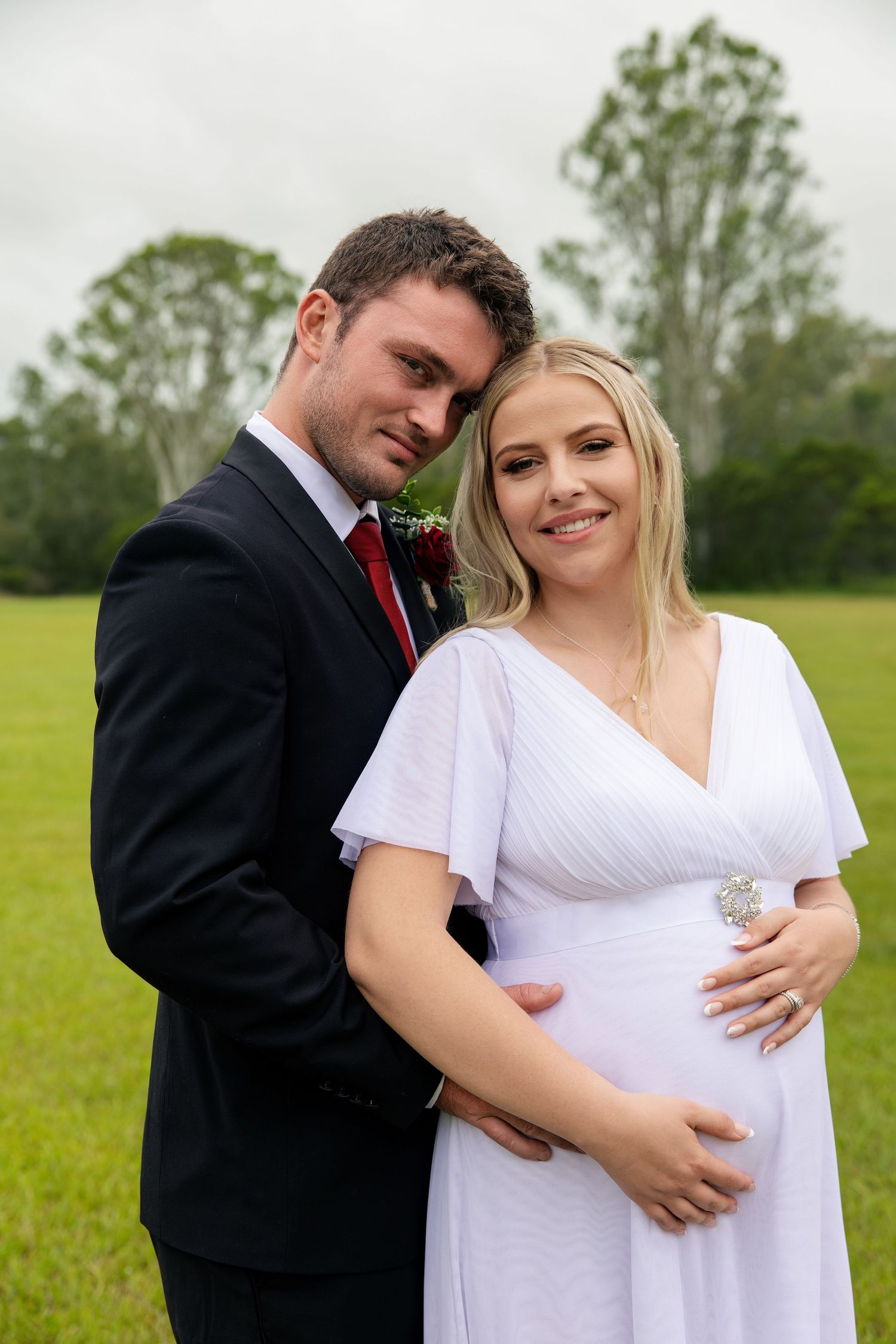 A couple in formal attire poses lovingly in a green field during a maternity photoshoot.