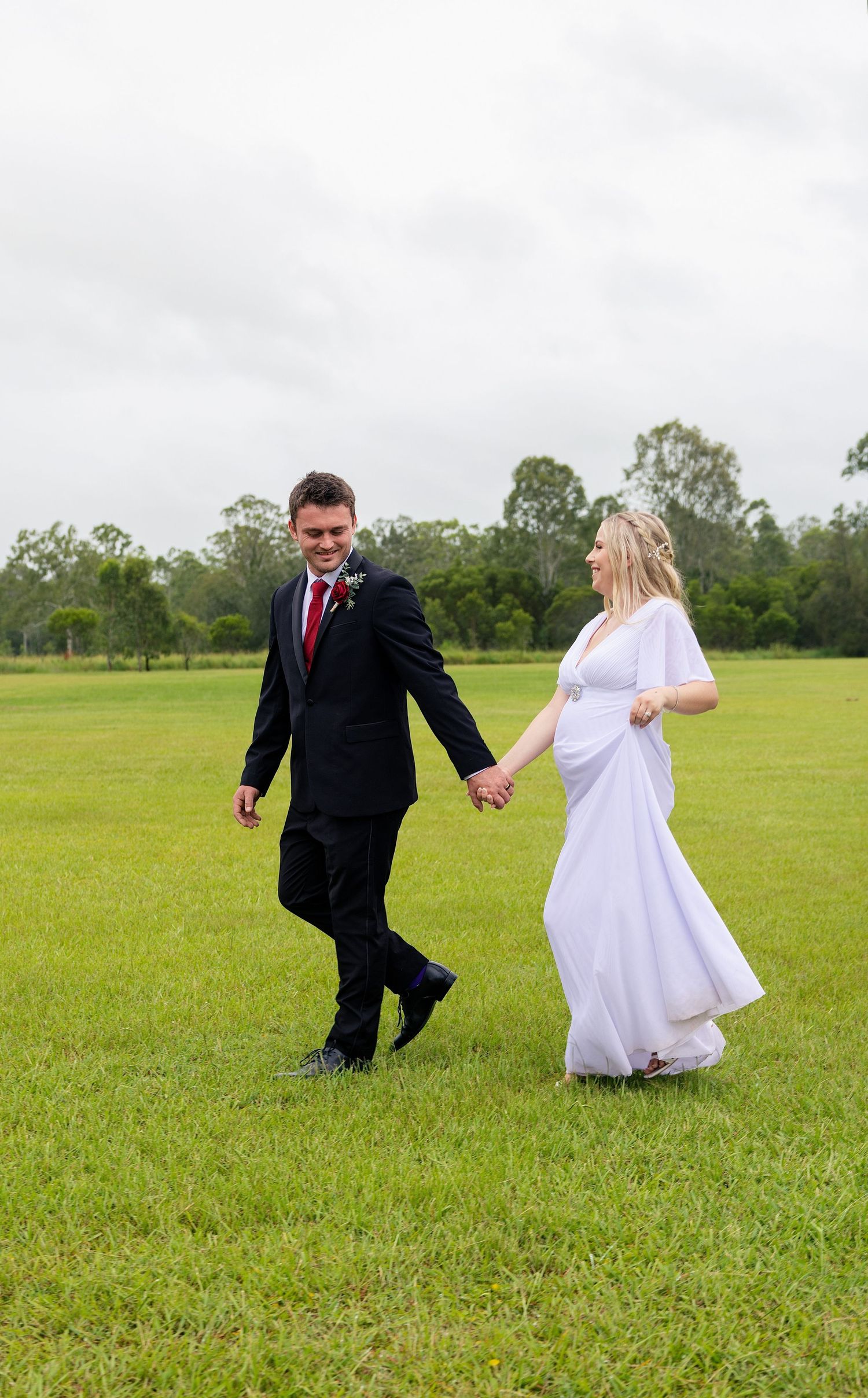 A couple in wedding attire playfully runs through a lush green field on their special day.