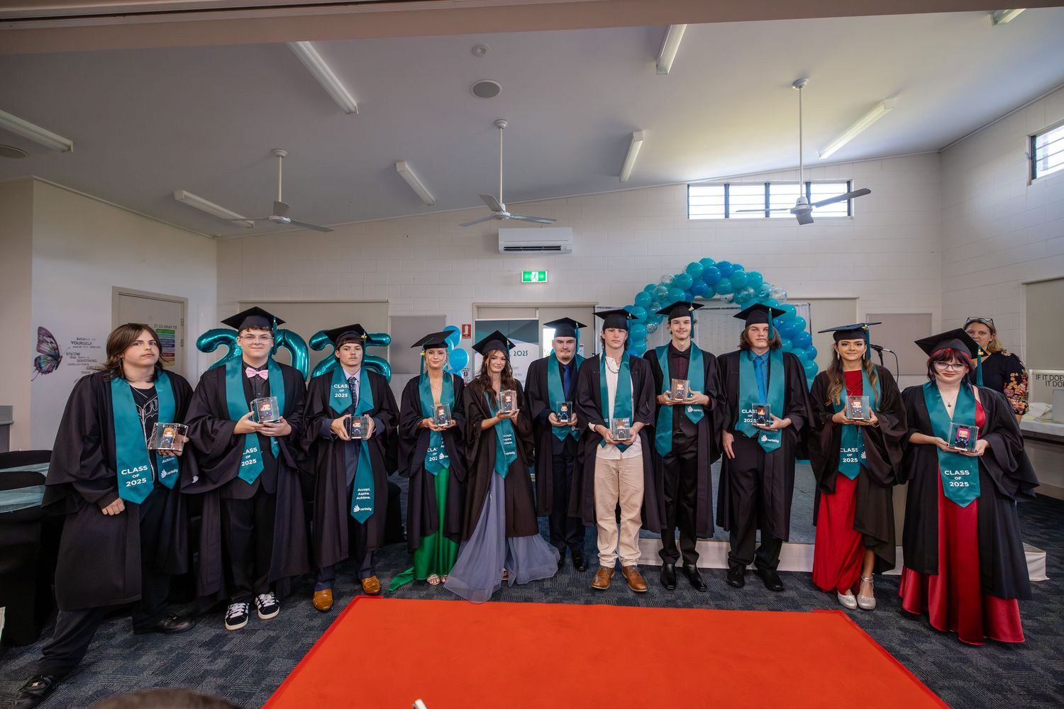 Group of graduates in black and teal academic gowns posing together at a graduation ceremony on a red carpet.