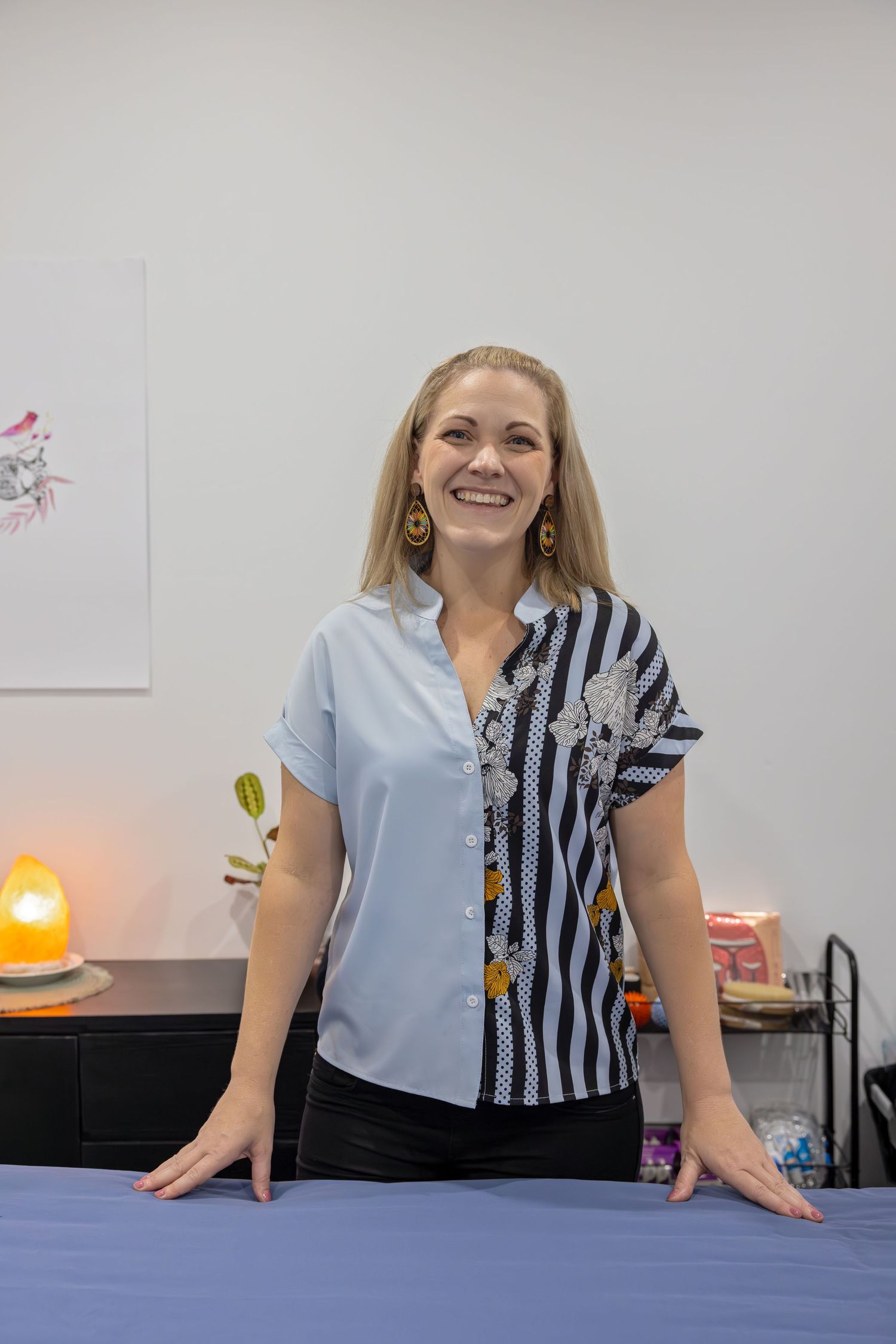 Smiling blonde woman in a striped and floral blouse standing confidently in a bright, welcoming spa room.