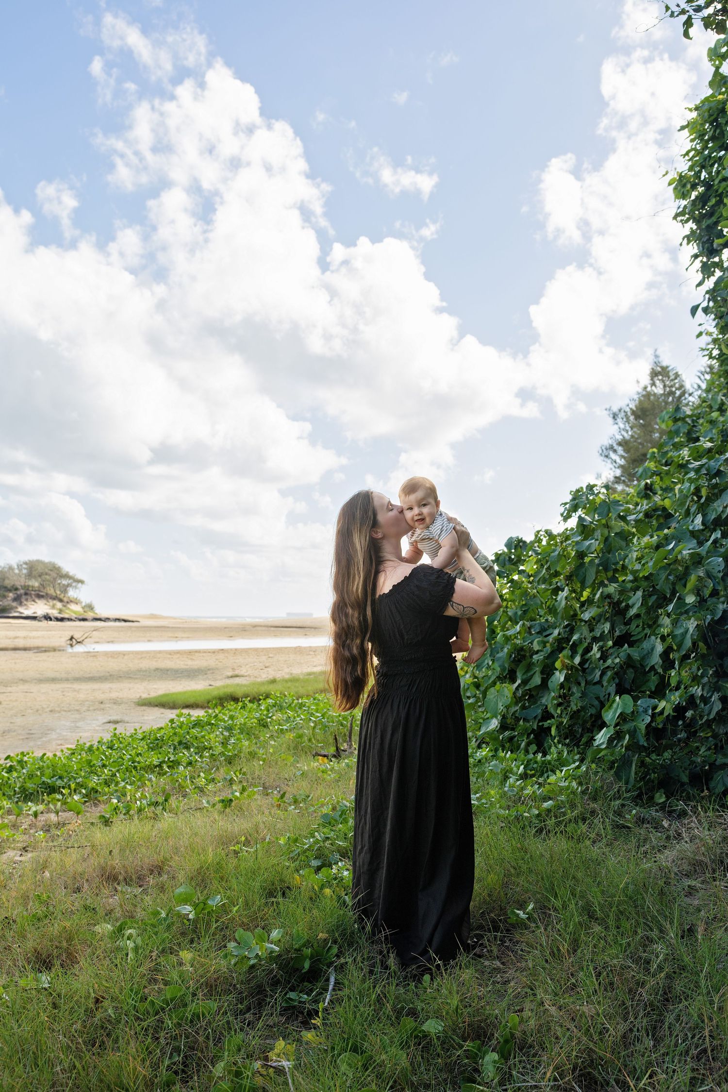 A couple in black attire shares an intimate moment in a grassy field with trees and cloudy skies in the background.
