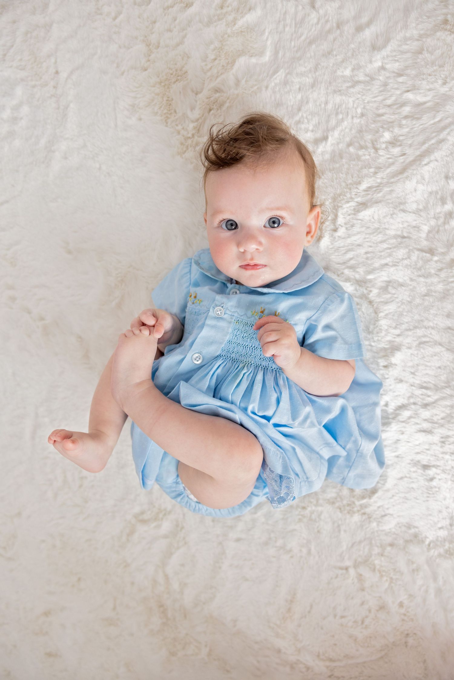 Baby wearing light blue dress lies on white textured blanket in series of four studio portraits.
