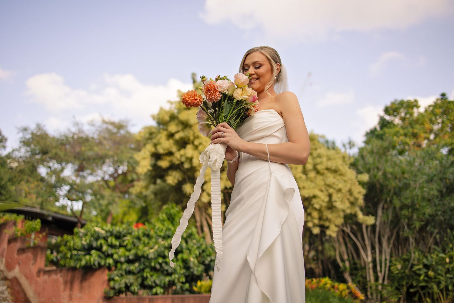 Bride in a white strapless wedding dress holds a vibrant sunflower bouquet in an outdoor garden setting.