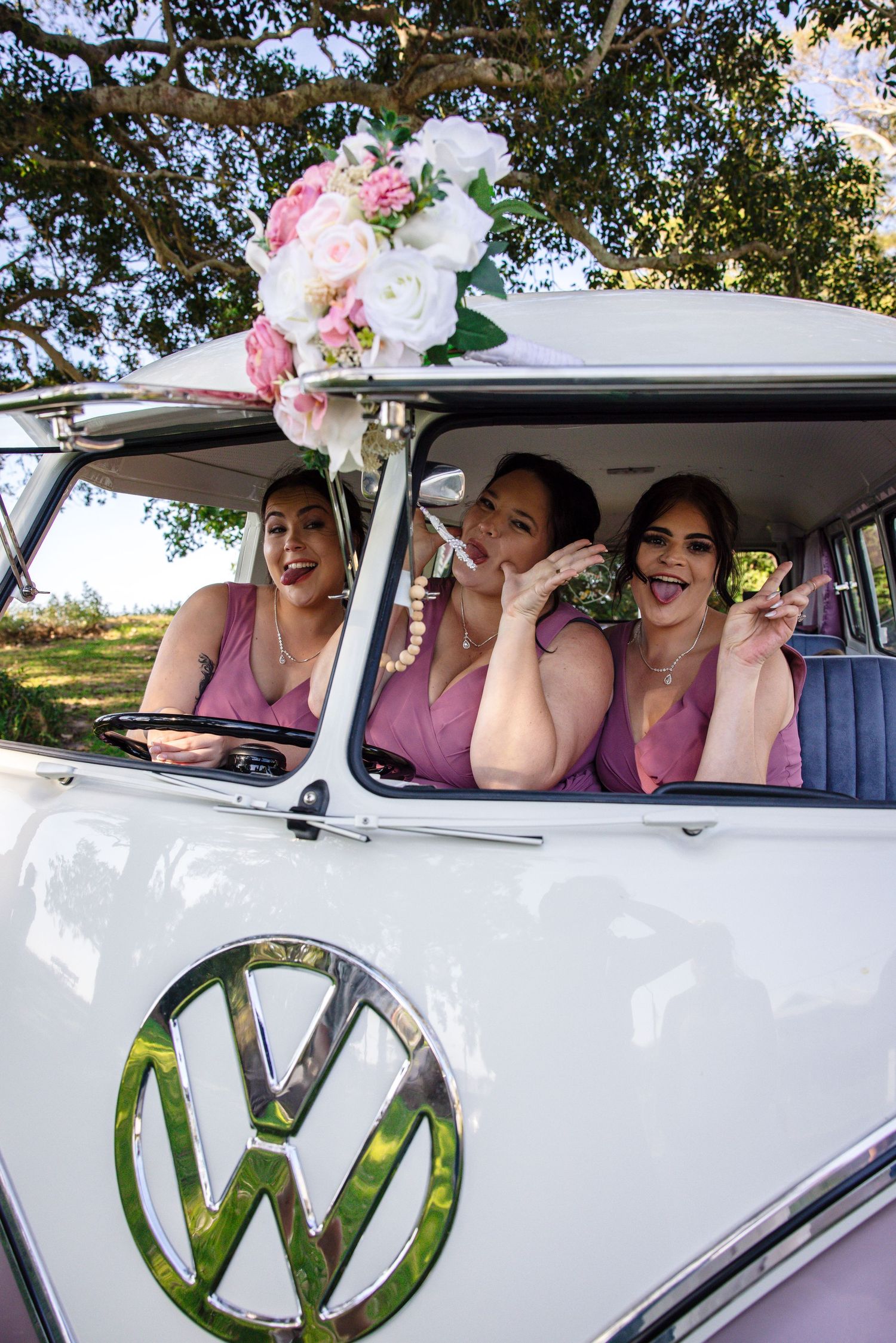 Three people peek out of a white vintage Volkswagen van window with floral decorations and visible VW logo.
