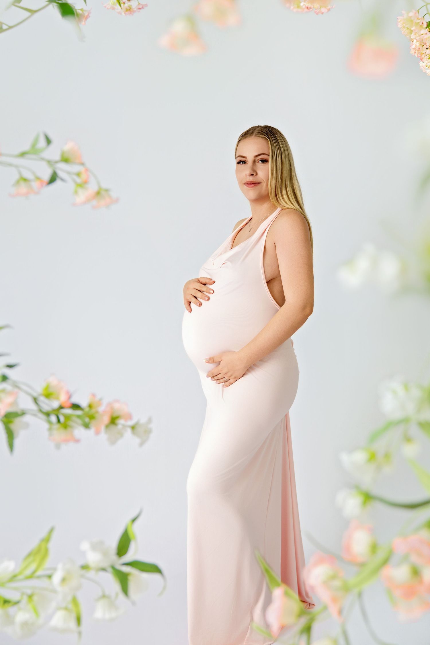 Pregnant woman in pink dress cradling baby bump surrounded by delicate flowers in a soft studio setting.
