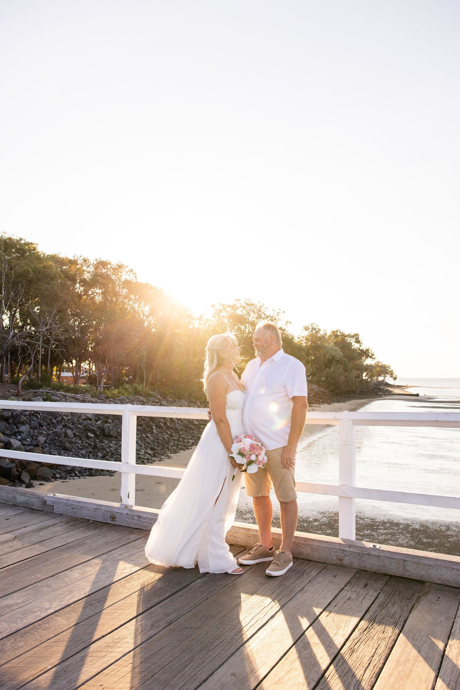 A couple shares a romantic moment on a wooden boardwalk at sunset with lens flare and white railing visible.