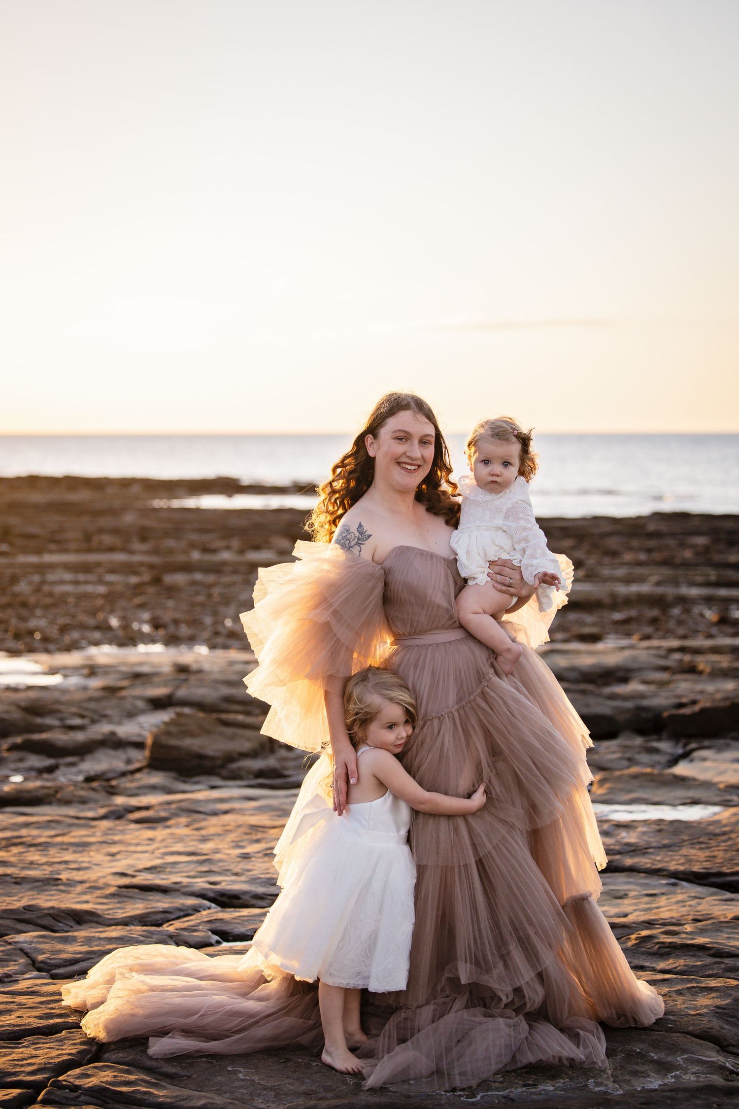 A tender family moment captured at sunset on rocky coastal rocks with flowing dresses in neutral tones.