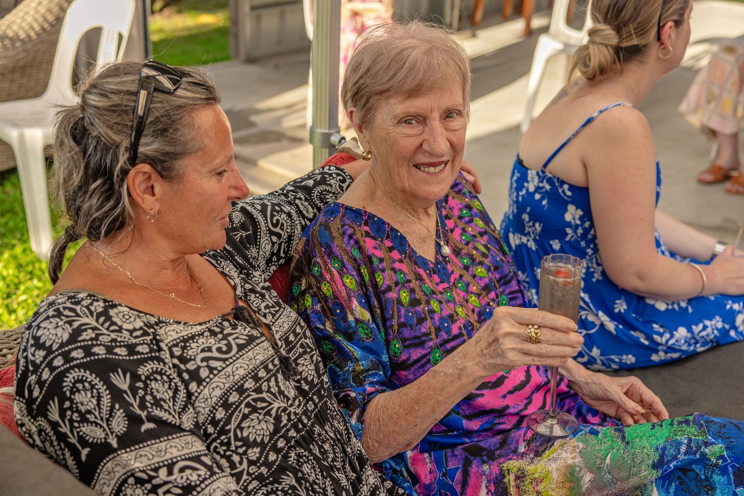 Two elderly women smiling and chatting outdoors at a sunny social gathering, one holding a drink.