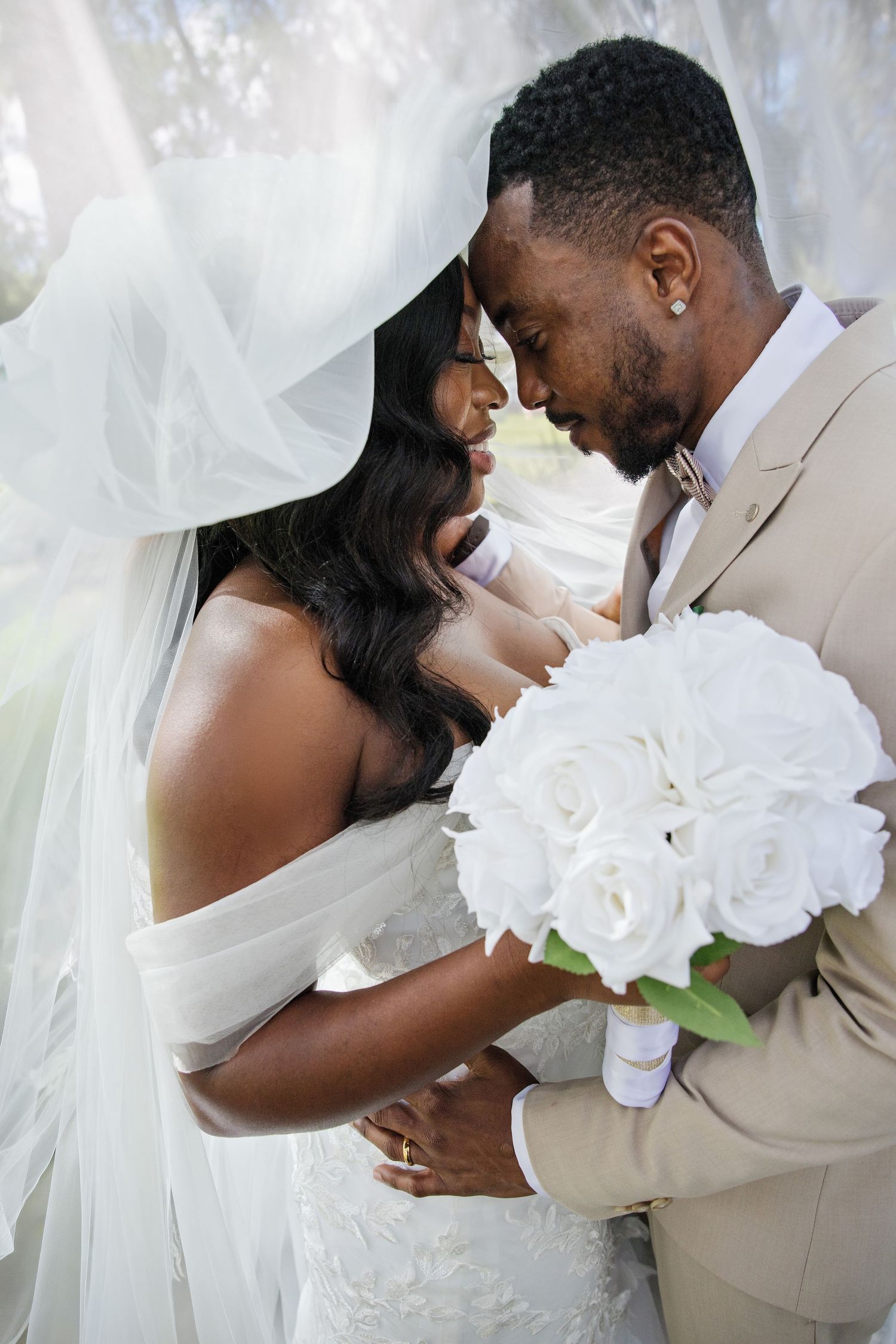 A Black bride and groom share an intimate moment under a flowing veil, holding white roses bouquet.