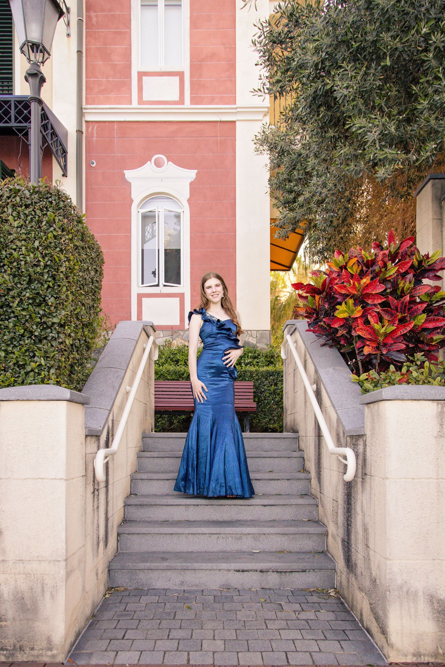 A person in a blue dress stands on concrete stairs with ornate stone railings and colorful plants in the background.