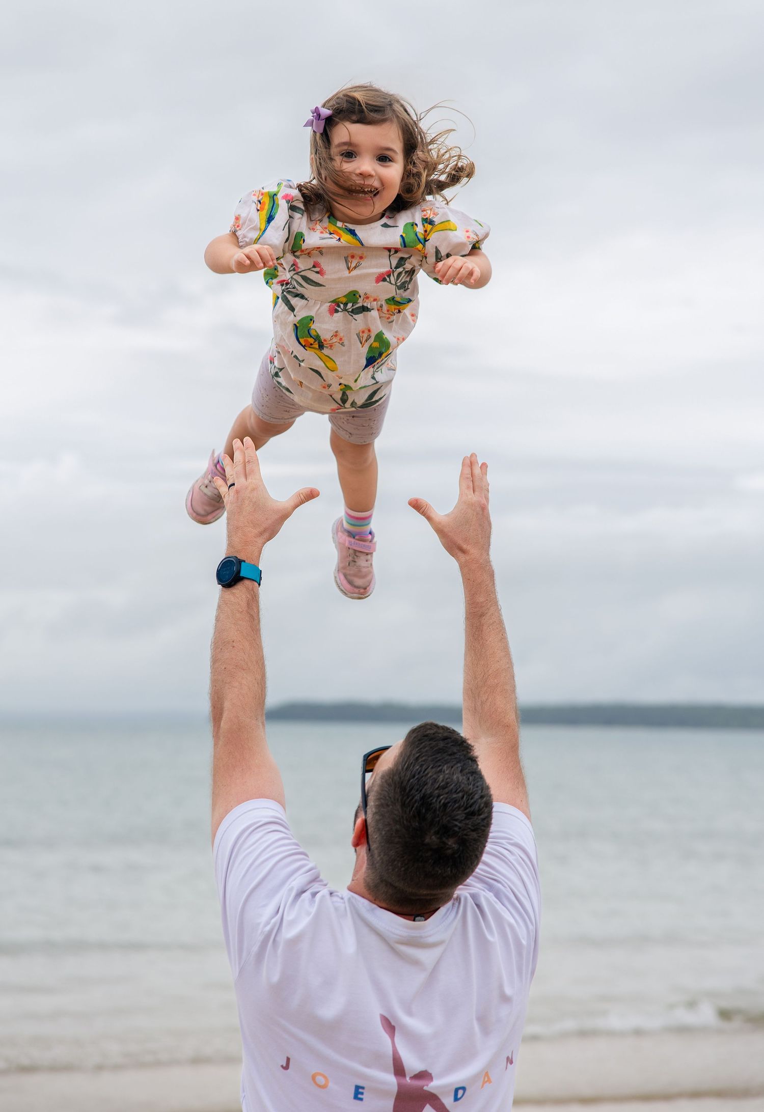A joyful father tosses his laughing young daughter into the air on a beach with an overcast sky behind them.