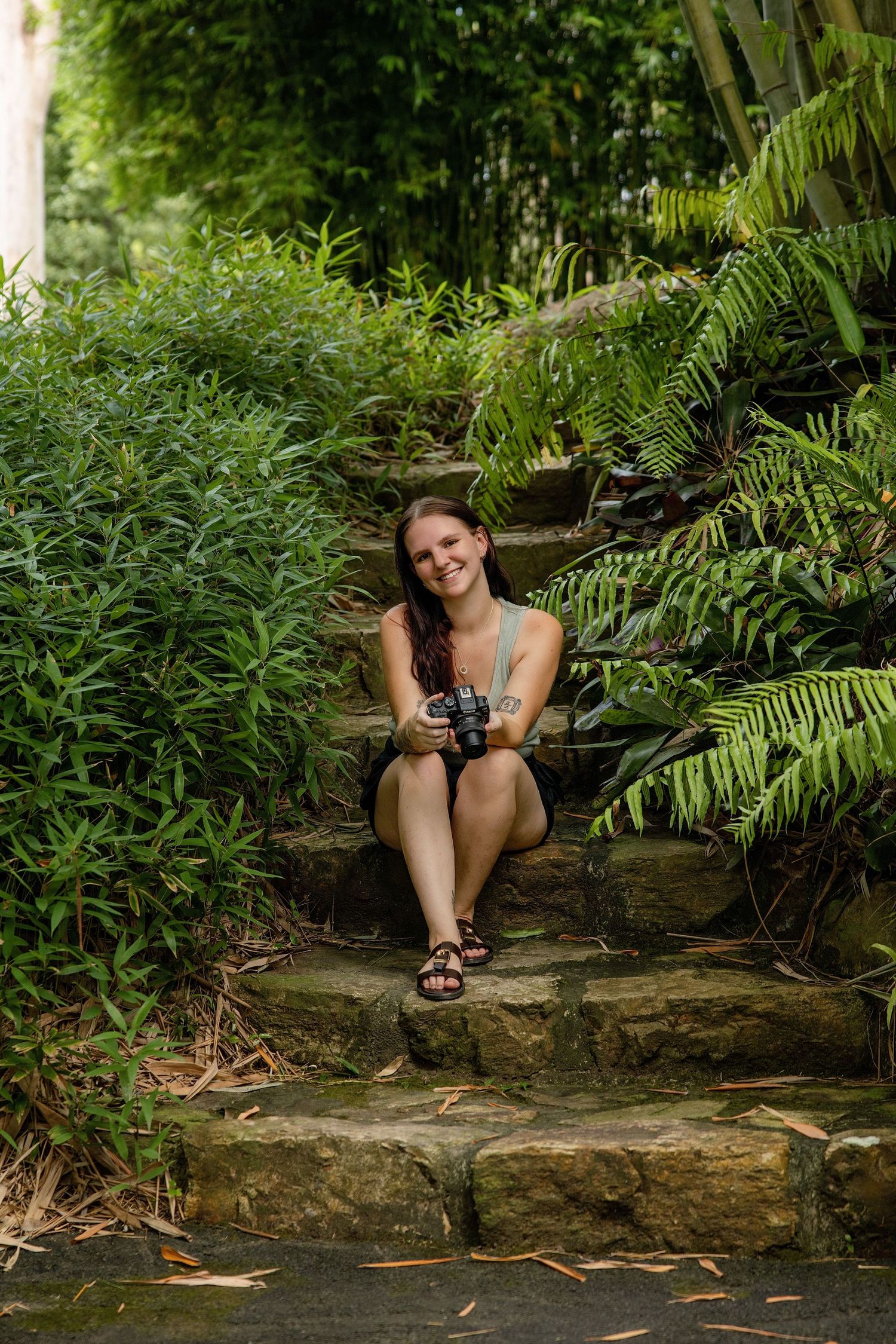 A person sits and smiles while relaxing on rocks surrounded by lush ferns and greenery in a natural setting.