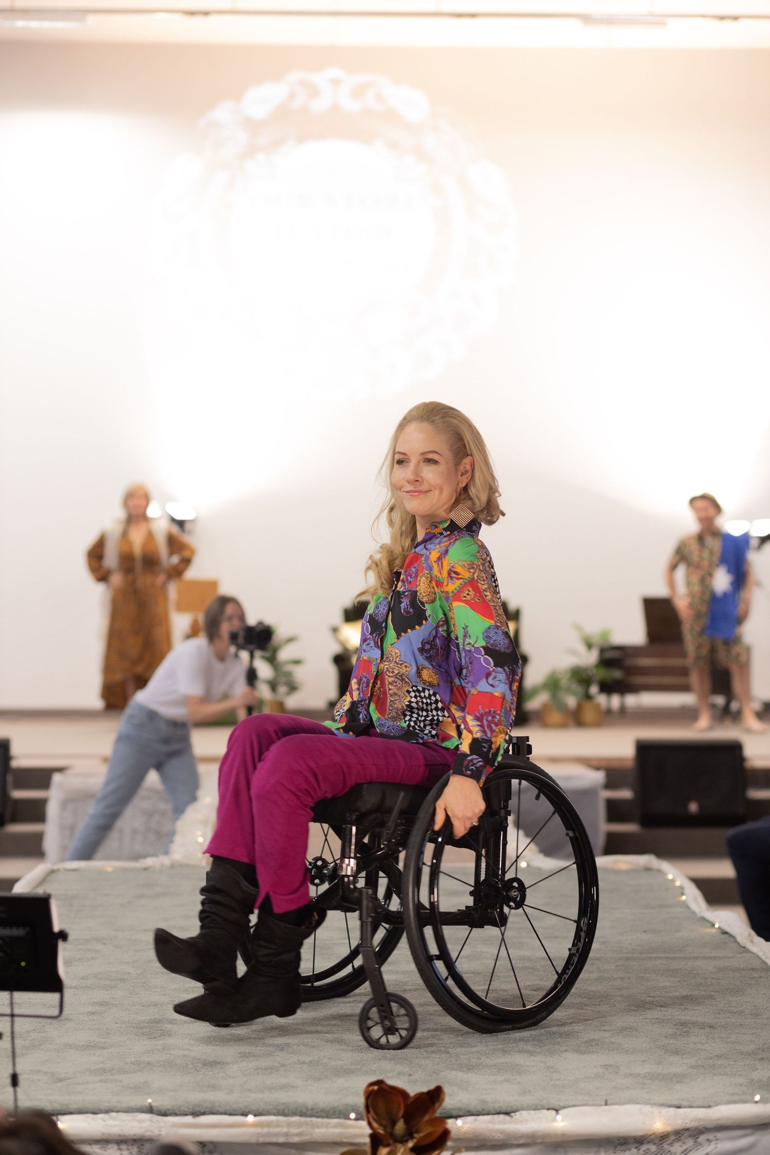 Young woman in colorful jacket and pink pants poses confidently in wheelchair on fashion runway.