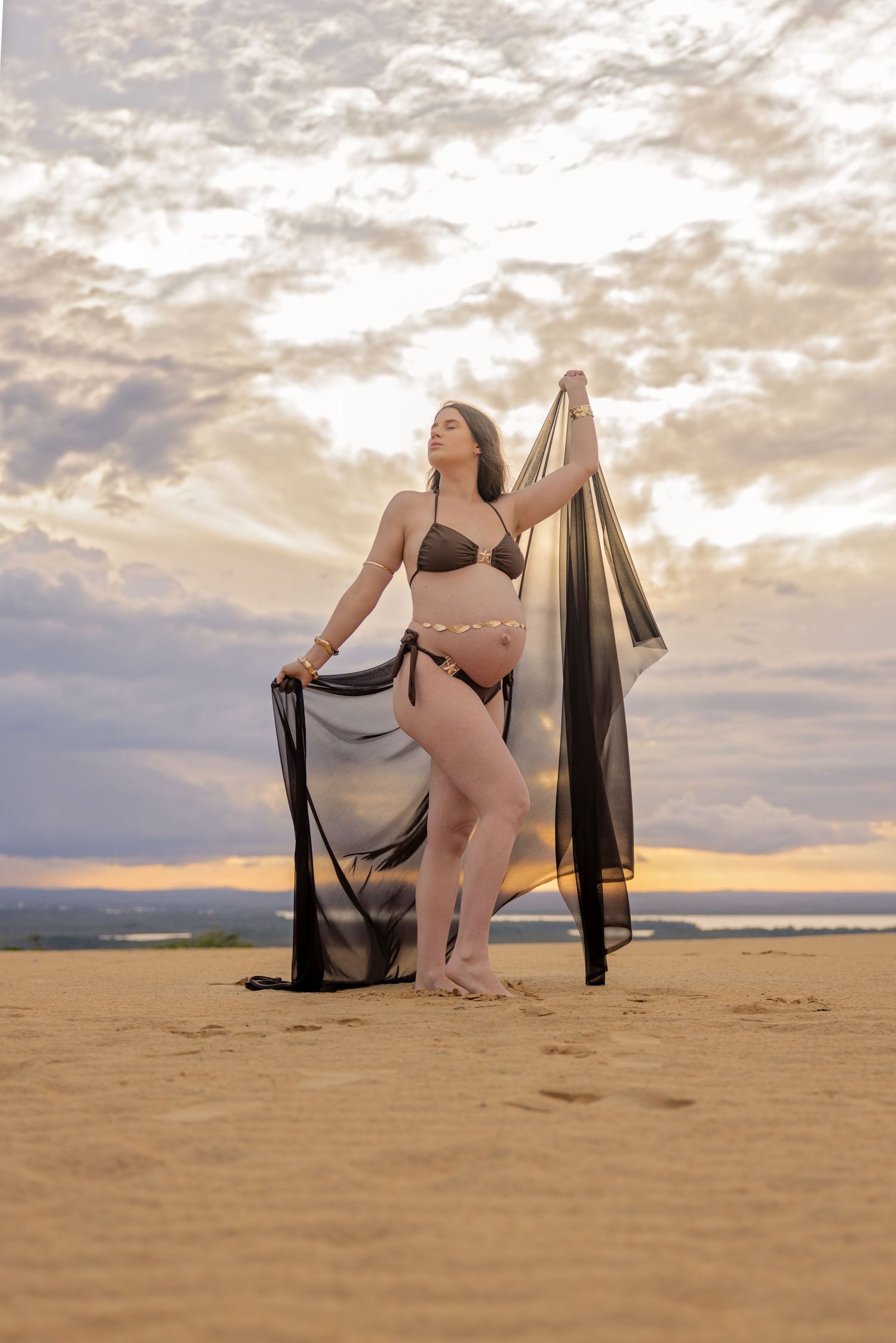 A figure in bikini poses with flowing black fabric on a beach during golden sunset with dramatic cloudy sky.
