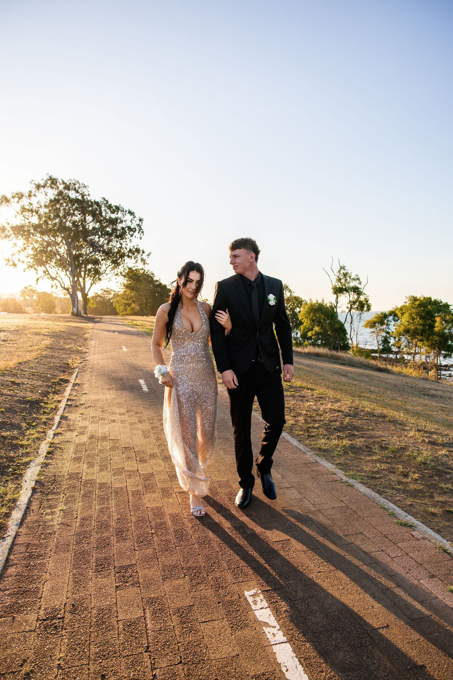 Couple in formal attire walking on a brick path at sunset, woman in sequin gown and man in black suit.