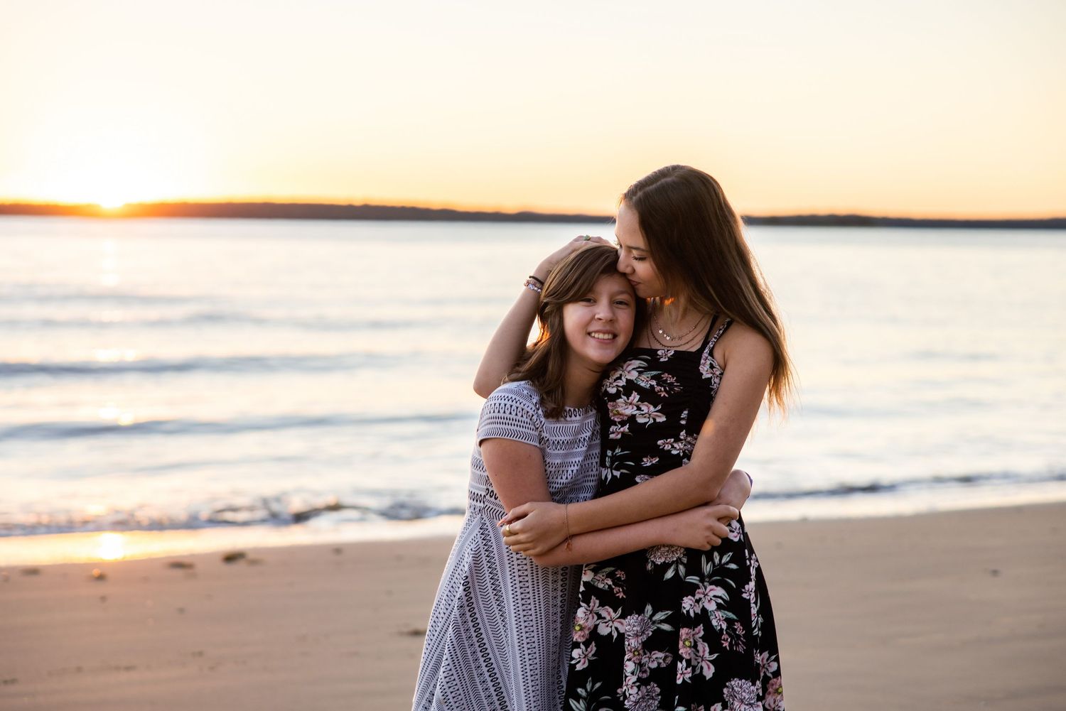 Two girls embracing and smiling on a sandy beach at golden hour sunset with calm ocean waves behind them.