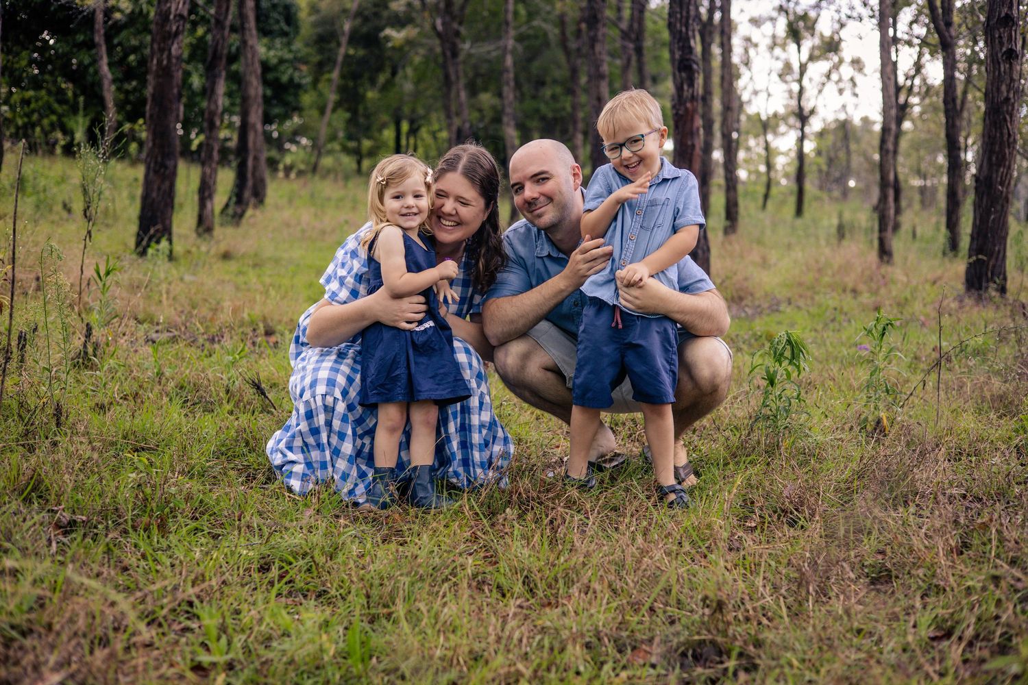 A loving family poses together outdoors in a wooded area with pine trees and green grass at sunset.