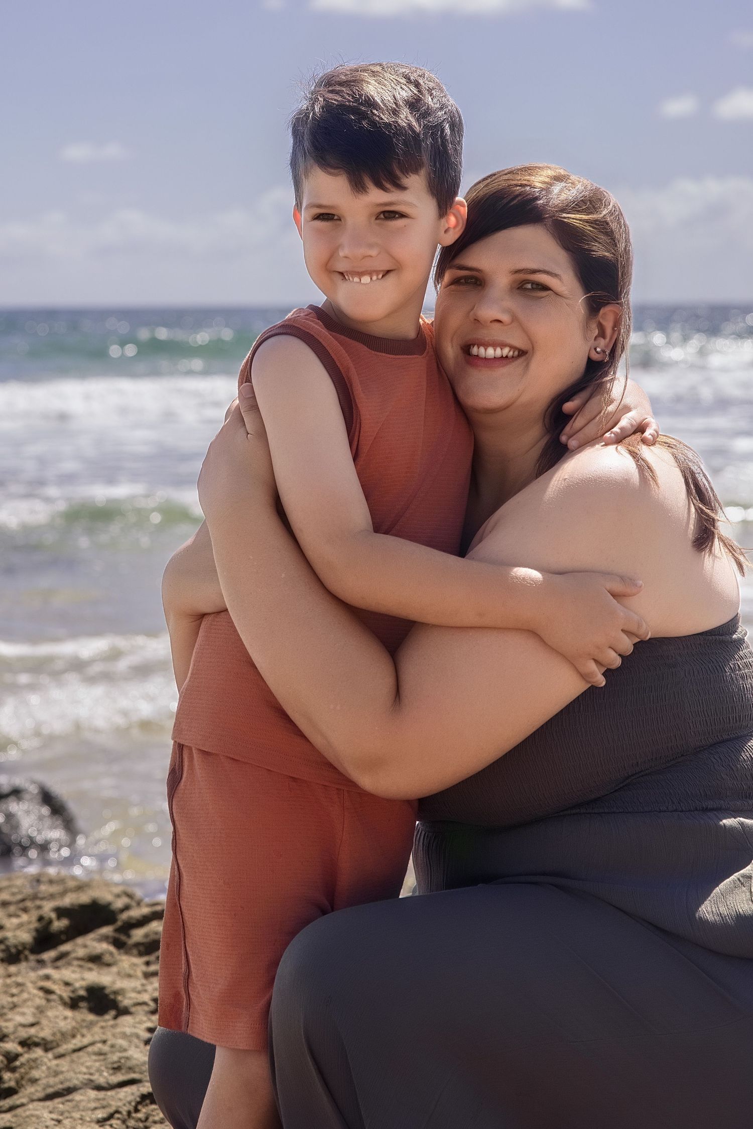 A parent and child embrace while sitting on rocky beach with ocean waves in the background on a sunny day.