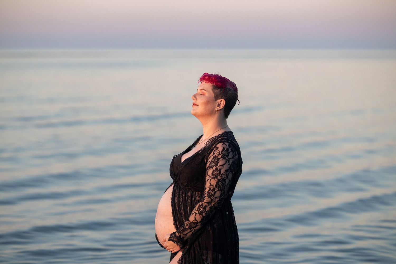 Pregnant woman in black lace dress holding belly by calm ocean at sunset with pink hair.