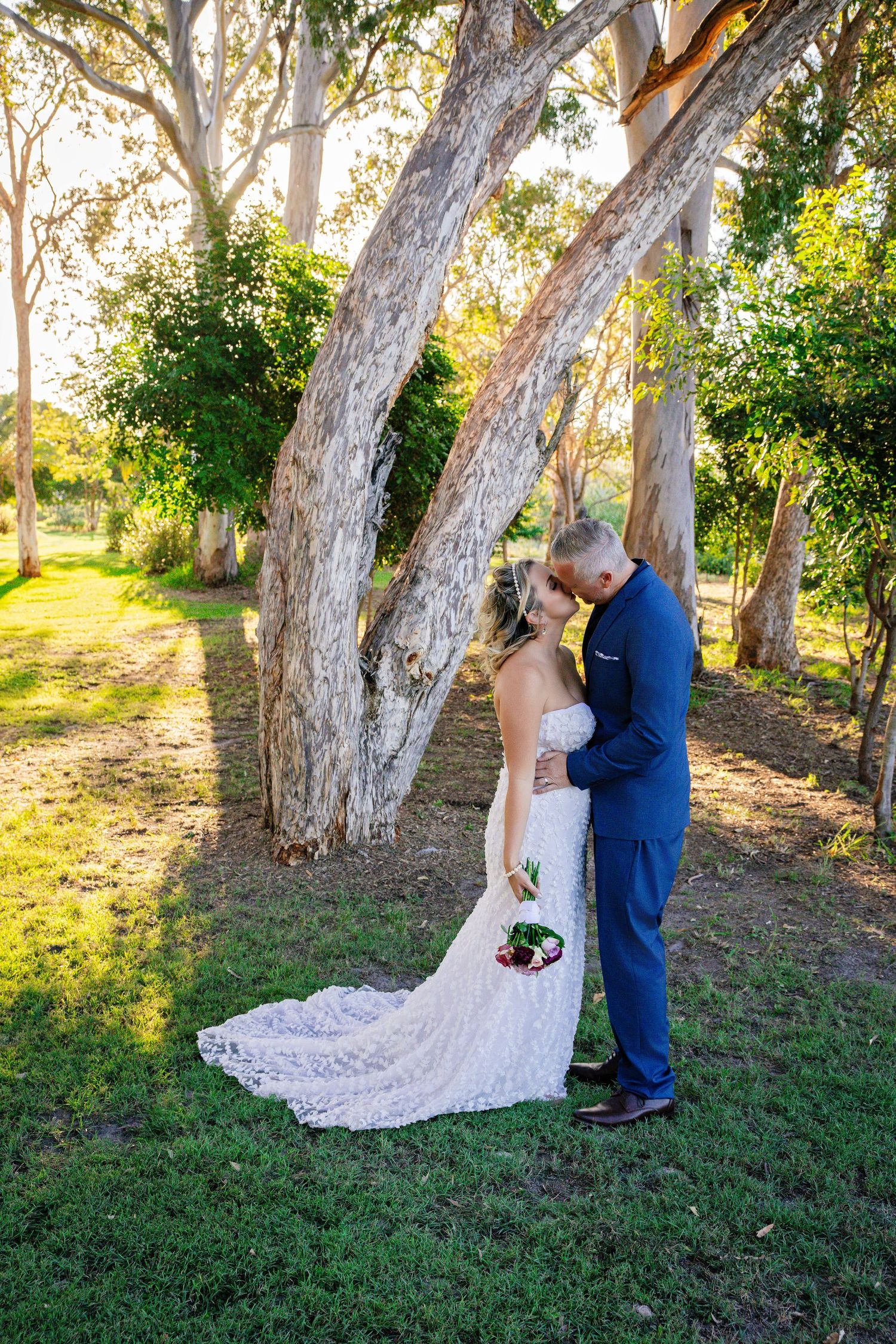 Bride and groom share a romantic kiss under eucalyptus trees, bride in lace gown holding bouquet, groom in navy suit.