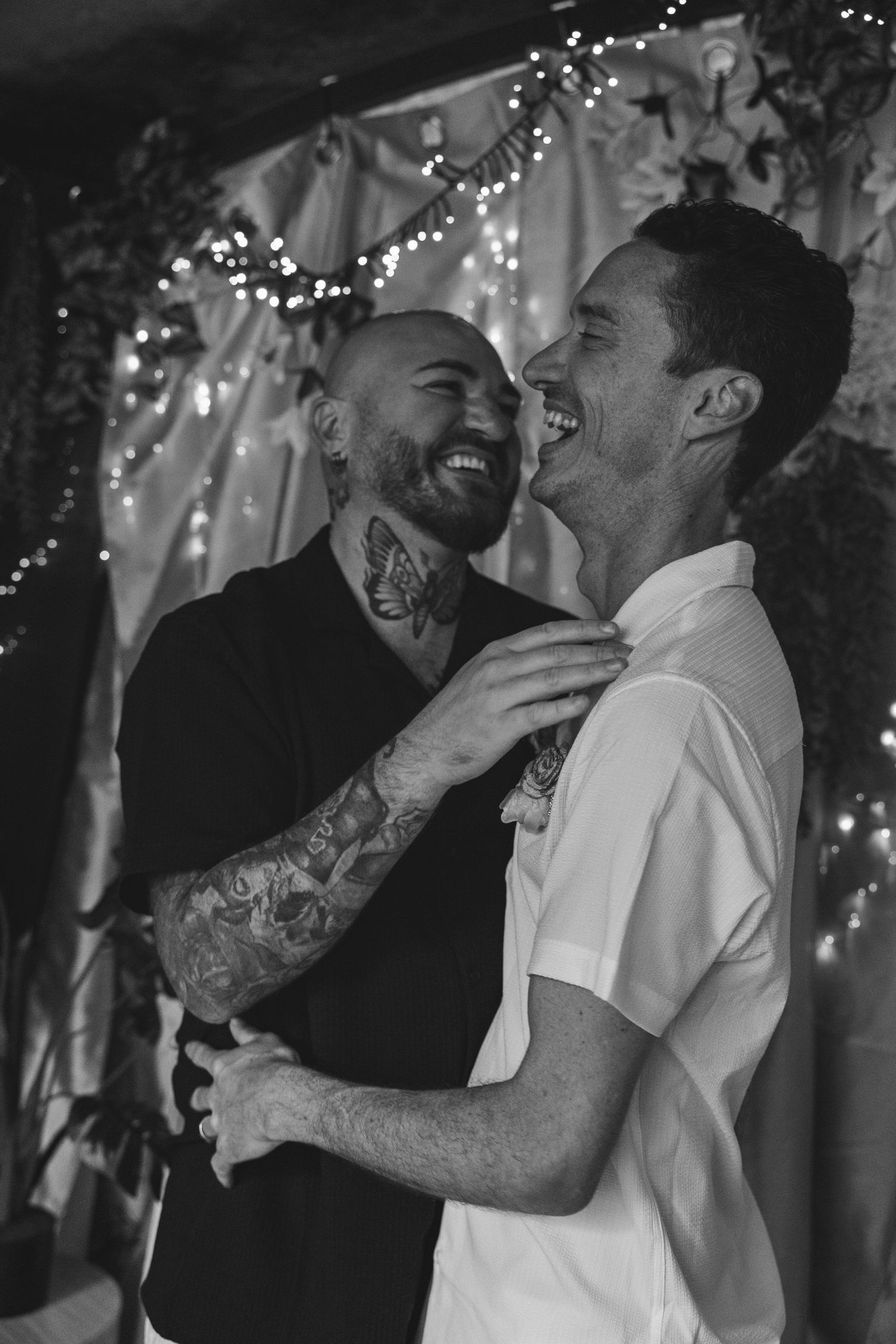 A loving black and white moment captured of two people laughing and embracing under decorative string lights.