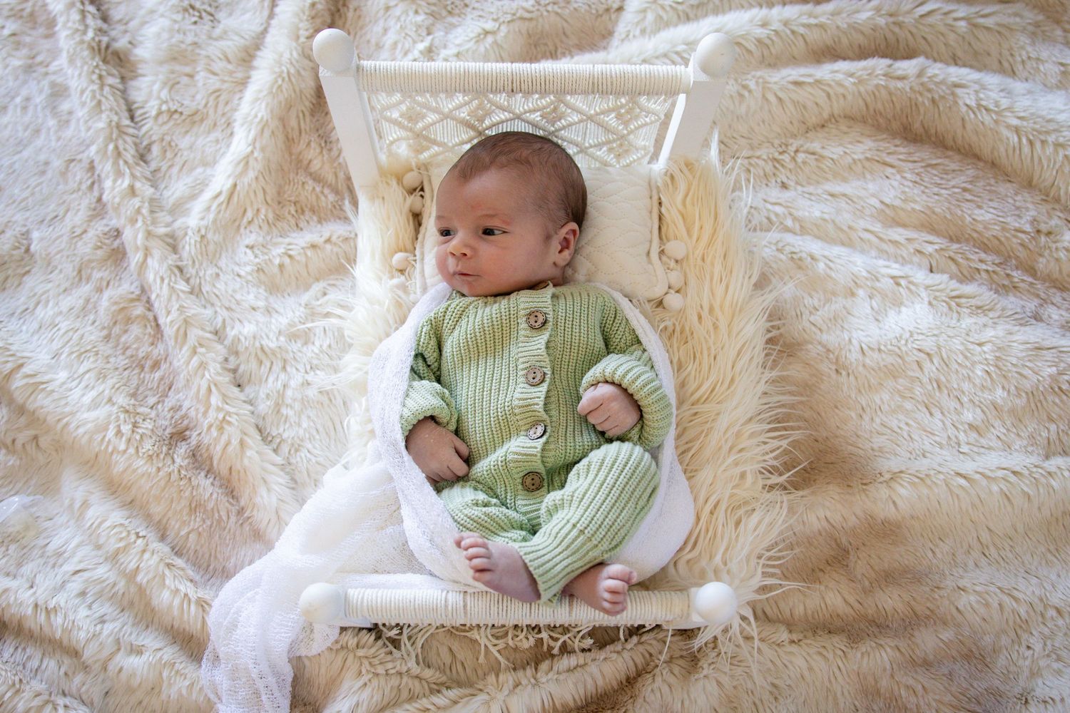 A baby in a mint green knitted romper lies on a fluffy white blanket looking peaceful.
