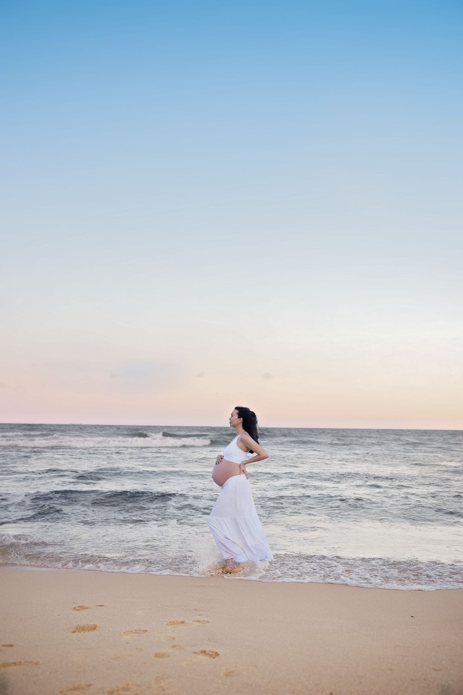 Person in white dress twirling joyfully on a sandy beach at sunset with ocean waves in the background.