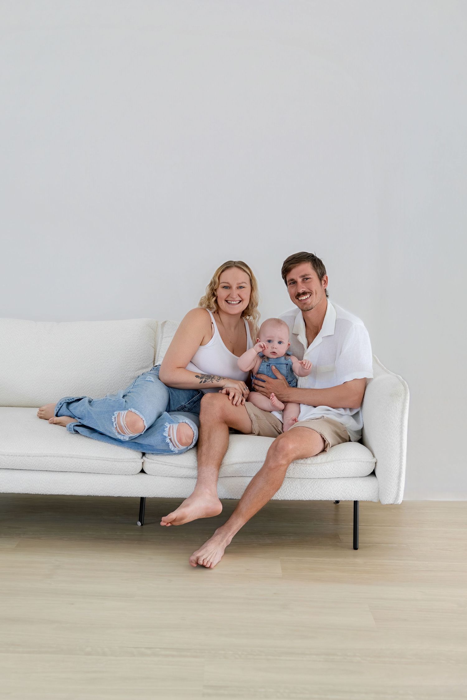 A young couple sits on a white sofa smiling, holding their infant baby dressed in blue denim.