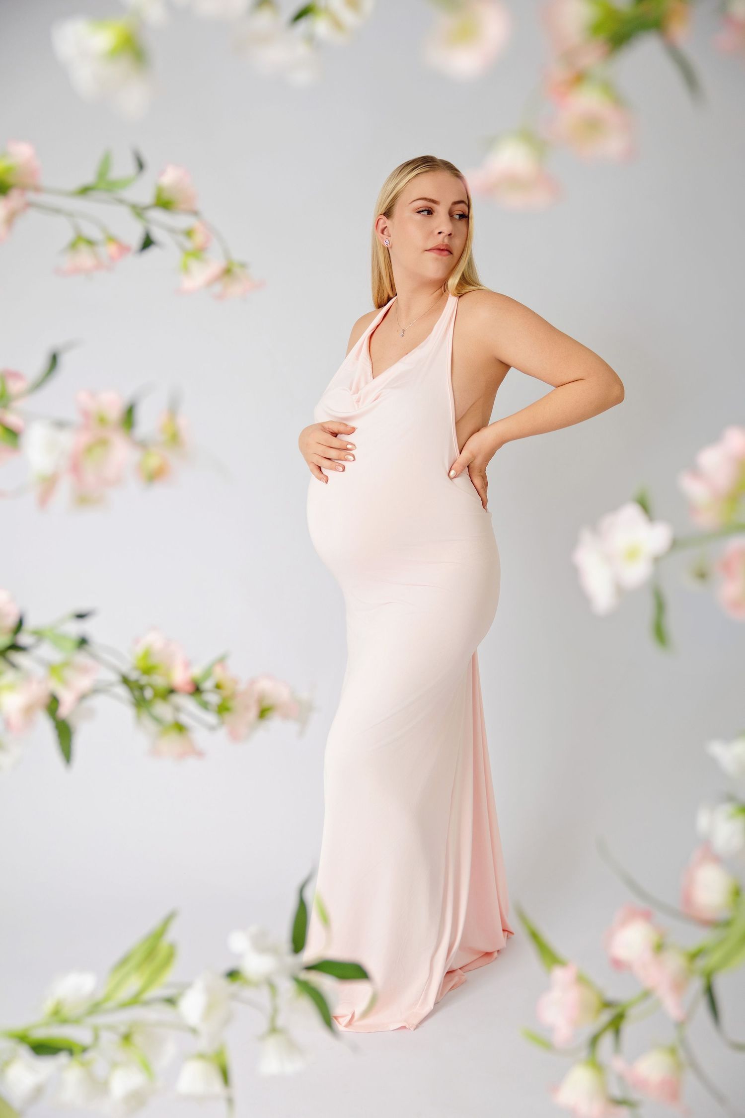 Pregnant woman in pink dress cradling baby bump surrounded by pink flowers.