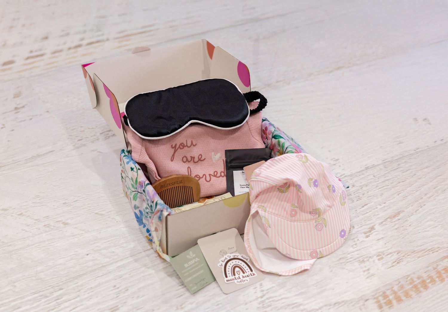 Pink gift box set with black sleep mask, 'you are loved' pouch, and pastel accessories on white wood.