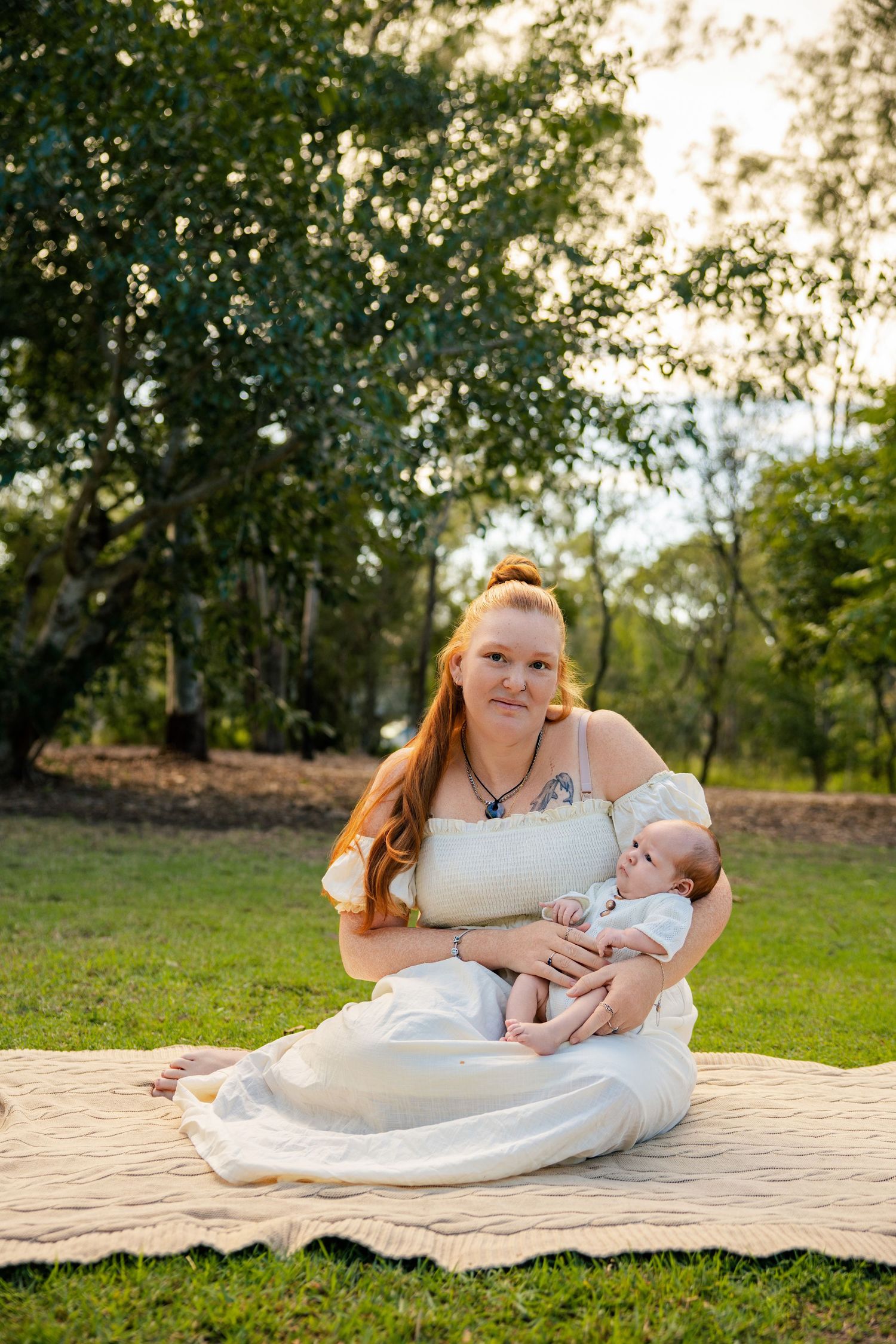 A young mother with red hair sits on a blanket in a park, cradling her newborn baby in a white dress.