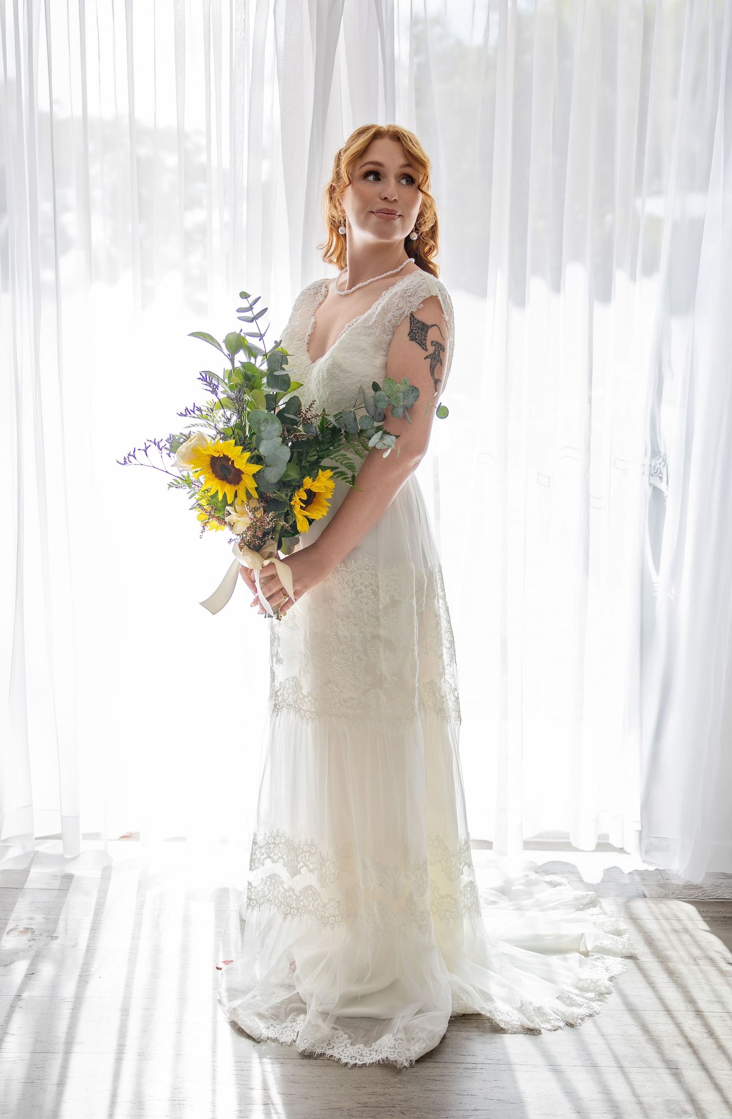 Bride in white lace gown holding sunflower bouquet stands before sheer curtains, showcasing tattoos and elegant updo.