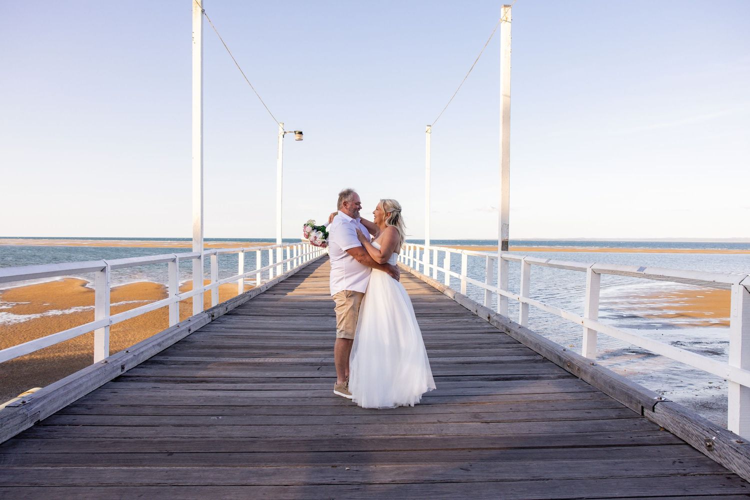 Wedding party walking along white wooden pier with ocean and blue sky in background.