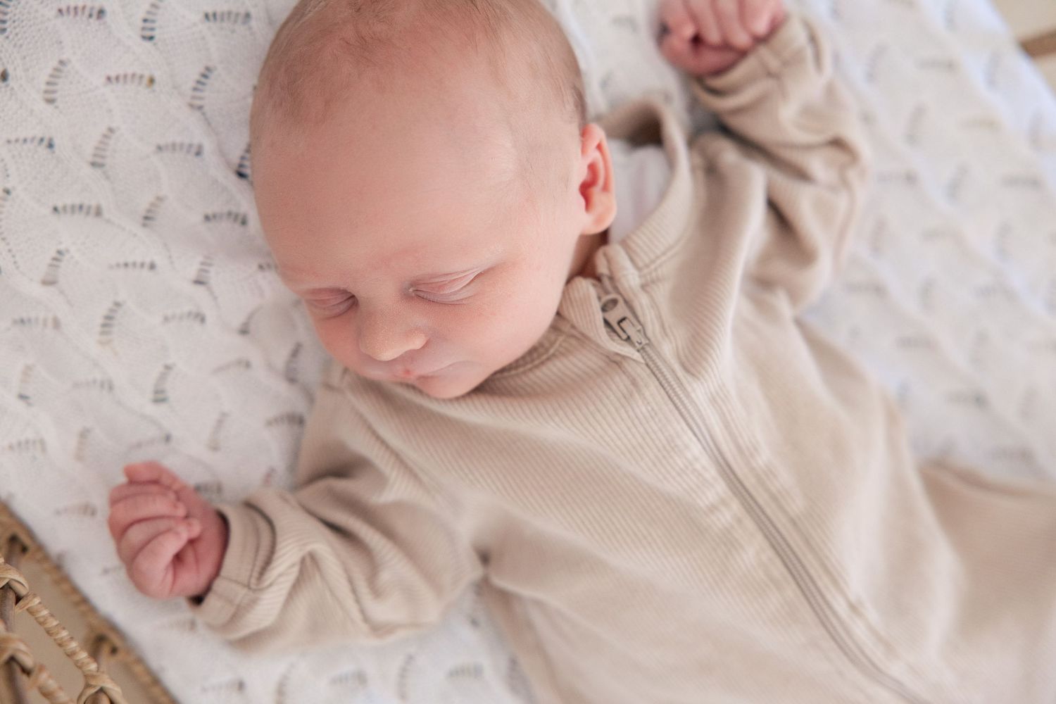 Newborn baby sleeping peacefully in a beige zip-up onesie on a white patterned blanket.