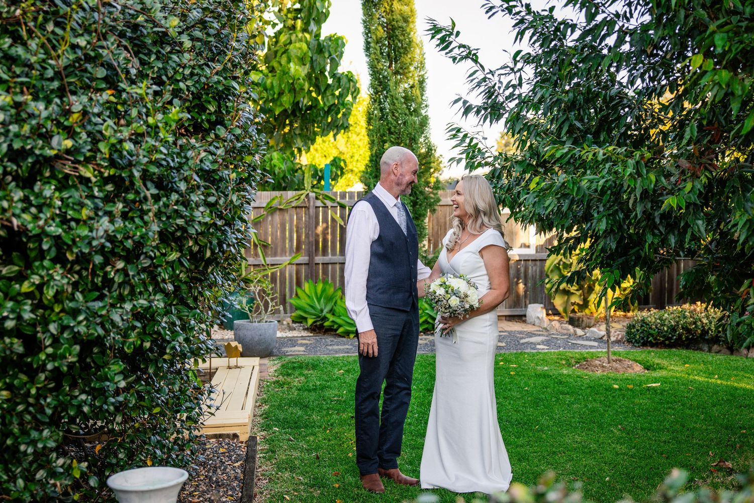 Couple in wedding attire shares a romantic moment in a lush garden setting at sunset.