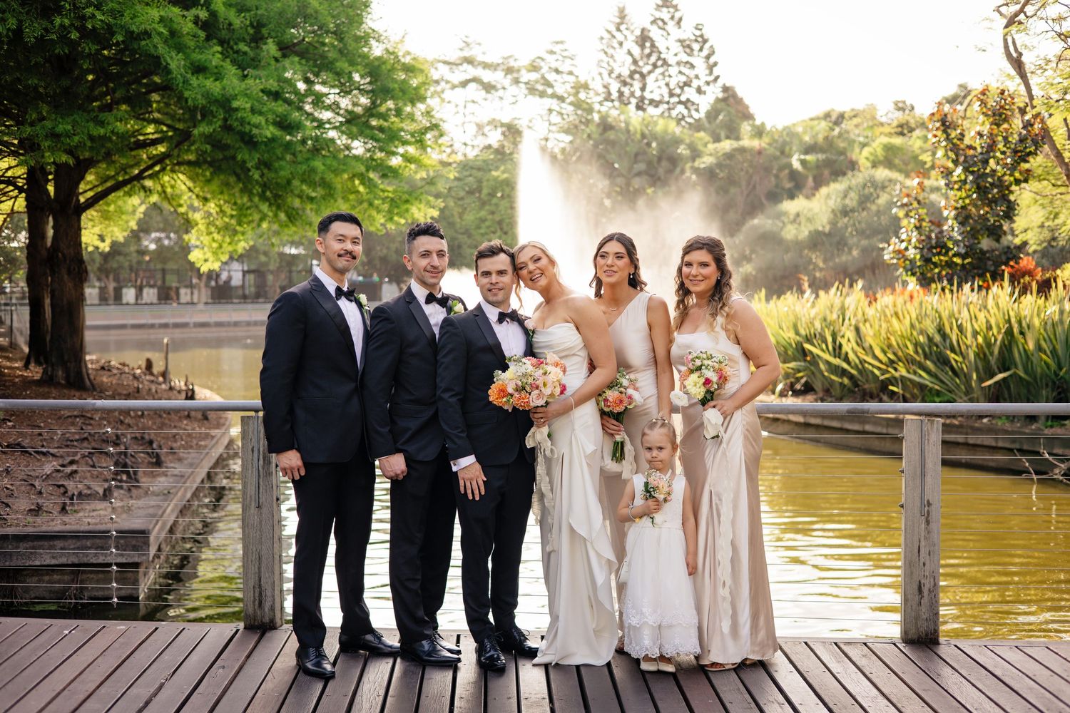 Wedding party poses together on rustic wooden deck by serene lake during golden hour sunset.
